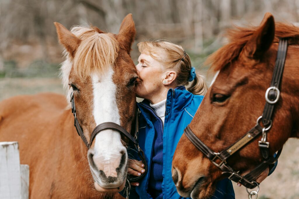 A woman lovingly kisses a horse on a sunny day, surrounded by nature.